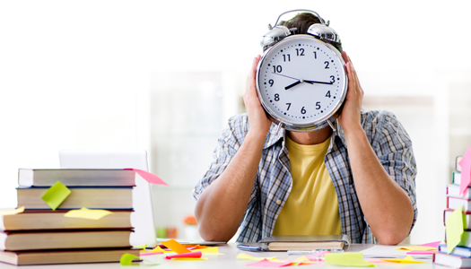 student sitting next to stack of books and holding a large clock up to his face