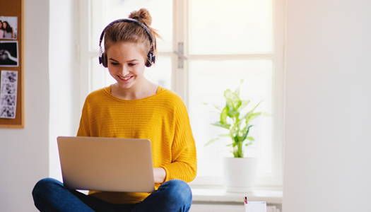 female sitting cross-legged wearing bright yellow sweater and headphones and typing on a laptop