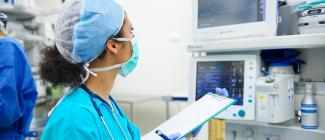 person wearing blue scrubs and face mask holding chart and looking at a monitor