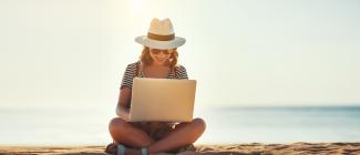 foreward facing female sitting crosslegged on beach with laptop in lap