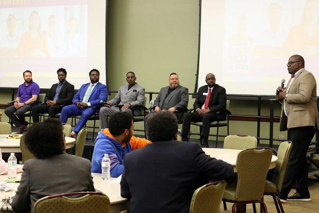 A panel discussion of 6 males with the panel moderator speaking